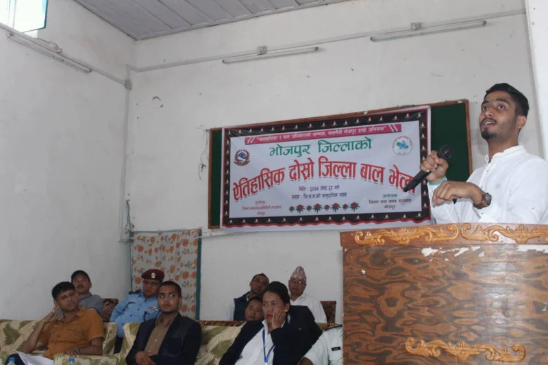 Dheeraj Joshi speaking at a lectern beneath a banner for the Bhojpur district children’s assembly, officials seated beside him.