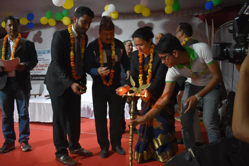 Guests in garlands lighting a ceremonial brass lamp on stage.