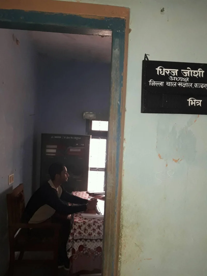 Dheeraj Joshi seated at a desk inside an office; a nameplate on the door frame bears his name and title.