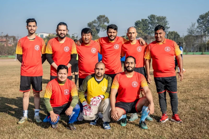 A football team in red and yellow jerseys posing on a grass pitch, Dheeraj Joshi crouching at front right.