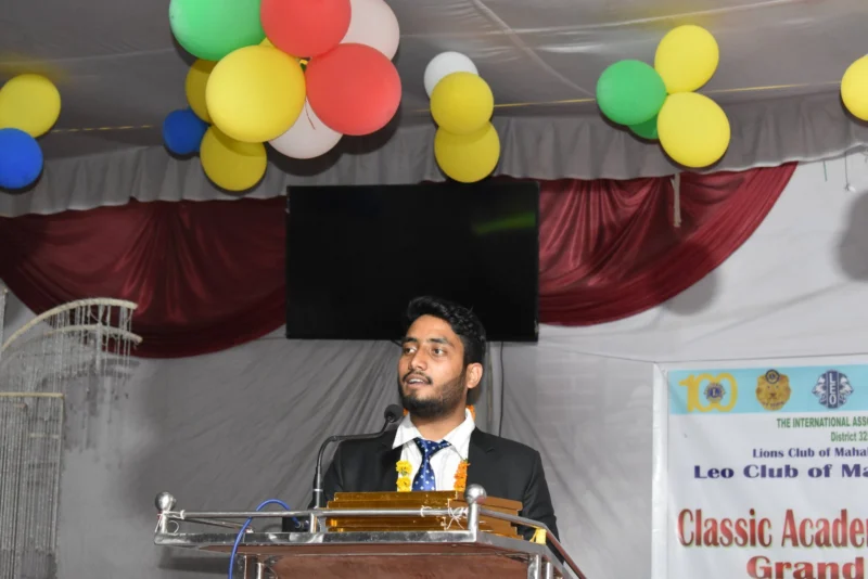 Dheeraj Joshi in a suit and garland speaking at a podium in front of a Leo Club banner.