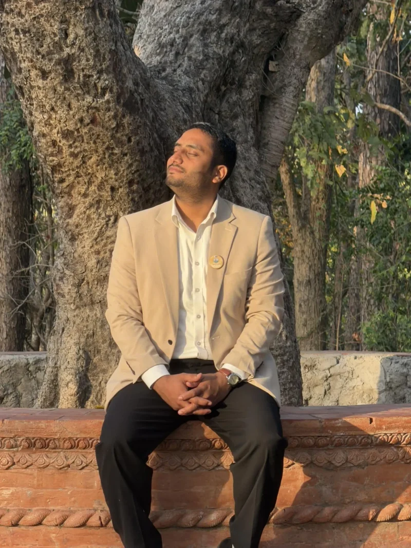 Dheeraj Joshi in a beige blazer, eyes closed in evening sunlight, seated on a carved brick platform beneath a large tree.