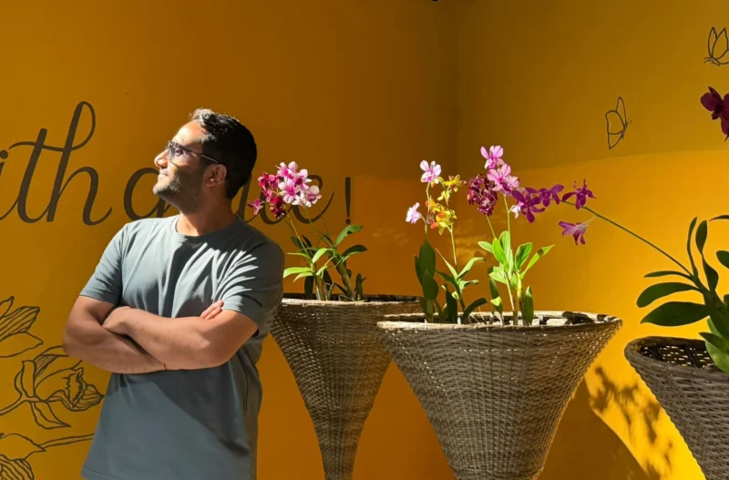 Dheeraj Joshi, arms folded and smiling, in sunlight against a yellow wall with orchids in woven planters.